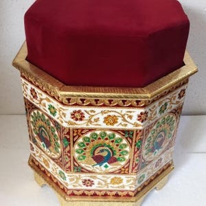 May include: An octagonal decorative stool with a plush, burgundy velvet top. The sides are white with gold trim and feature peacock designs in red, green, and blue. The base and trim are gold.