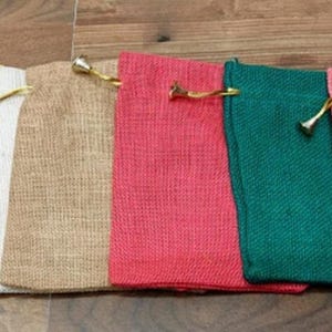 Può includere: Sei sacchetti in iuta con coulisse in fila, in giallo, bianco, beige, rosso, verde e rosa. Ogni sacchetto ha un'estremità metallica dorata sul cordoncino. I sacchetti sono disposti uno accanto all'altro su una superficie di legno.