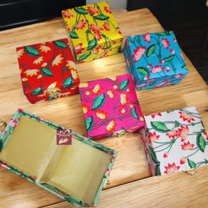 Può includere: Cinque piccole scatole quadrate con motivi floreali. Le scatole sono in legno e hanno un coperchio incernierato. I motivi sono fiori di loto e foglie in vari colori, tra cui rosso, giallo, blu, rosa e bianco.