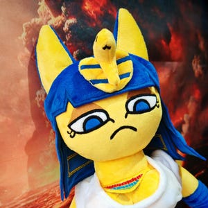May include: A plush toy featuring a yellow cat-like character with blue accents. The character wears a blue headdress with a yellow snake and a white dress. The toy has large blue eyes and a slightly grumpy expression.