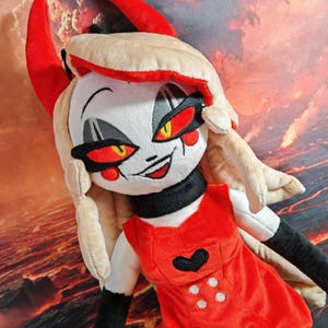 May include: Plush doll of a character with white skin, red eyes, and blonde hair. The doll is dressed in a red dress with a black heart and white buttons. It has black arms and a black choker. Red horns and a smiling expression complete the design.