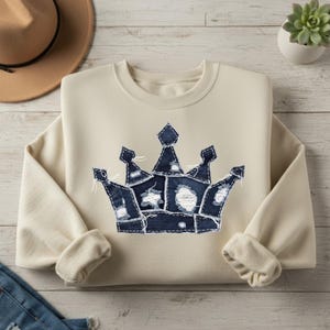 May include: Cream-colored sweatshirt featuring a large, distressed denim crown applique. The crown is made of various denim patches with white stitching and frayed edges. The sweatshirt has a crew neck and long sleeves with ribbed cuffs.