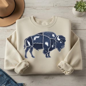 May include: Cream-colored sweatshirt featuring a denim patchwork bison design. The bison is outlined with white stitching, and the denim patches have distressed details. The sweatshirt has long sleeves with ribbed cuffs and a crew neck.