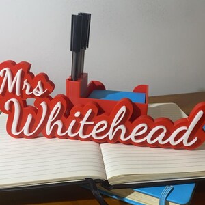 May include: Red and white 3D printed desk organiser with the name "Mrs. Whitehead" on it. The organiser has a pen holder and a space for sticky notes.