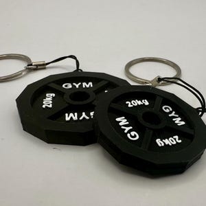 May include: Two black weight plate keychains with silver key rings. Each keychain features the text "GYM" and "20kg" in white lettering.