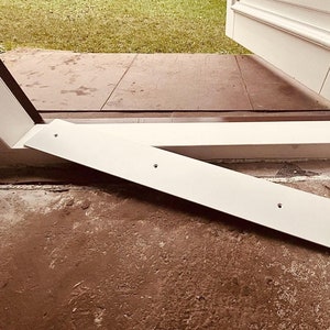 May include: White door frame and trim with a white threshold piece. The door is open, showing a view of the garden. The threshold has pre-drilled holes. The floor is a mix of concrete and stone.