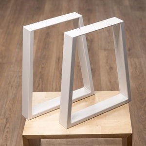 May include: Two white metal furniture legs with a trapezoidal shape. Each leg features a flat top mounting plate with pre-drilled holes for attachment. The legs have a smooth, matte white finish and a sturdy, rectangular tubular construction.