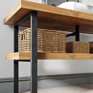 Può includere: Un mobile da bagno con un piano in legno marrone chiaro spesso e un ripiano inferiore sostenuto da una struttura in metallo nero. Due cesti in vimini intrecciato si trovano sul ripiano inferiore. Un lavabo bianco e un rubinetto nero sono sopra.