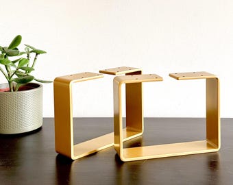 Könnte beinhalten: Drei goldene Möbelbeine aus Metall mit quadratischer Form und abgerundeter Oberseite. Die Beine sind in einer Reihe auf einer dunkelbraunen Oberfläche angeordnet.