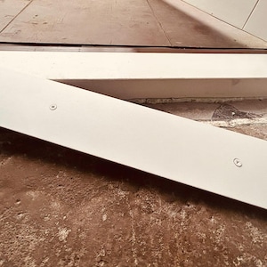 May include: A section of a white door frame with two visible screws. The frame is angled across a textured, reddish-brown surface. The top of the frame is visible, along with a section of the floor.