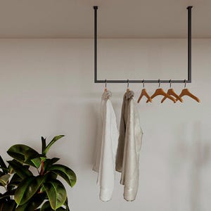 Könnte beinhalten: Eine schwarze Metall-Kleiderstange, die von der Decke abgehängt ist und Holzkleiderbügel mit Kleidungsstücken hält. Ein weißes Hemd und eine hellfarbene Jacke hängen an der Stange. Eine grüne Pflanze ist unten links zu sehen.