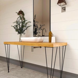 Può includere: Un mobile da bagno con un piano d'appoggio rettangolare in legno chiaro sostenuto da quattro gambe a forcina in metallo nero. Un lavabo bianco è posizionato sulla superficie con un rubinetto argentato. Uno specchio rettangolare con cornice nera.