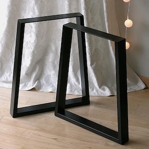 May include: Two black metal table legs with a trapezoidal frame design. Each leg features a sturdy, rectangular steel construction with a smooth matte finish, angled sides, and a flat base for stable support on flat surfaces.