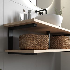 Peut inclure: Meuble-vasque de salle de bain moderne avec une étagère en bois clair et des supports en métal noir. Deux paniers en osier tressé sont posés sur l'étagère inférieure. Un lavabo blanc et un robinet sont visibles au-dessus.