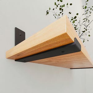 Op de afbeelding: Een lichtgekleurde houten plank gemonteerd aan een muur met twee matzwarte metalen beugels. De plank heeft een natuurlijke houtnerfstructuur en is aan de muur bevestigd met zichtbare schroeven op de verticale en horizontale delen van de beugels.