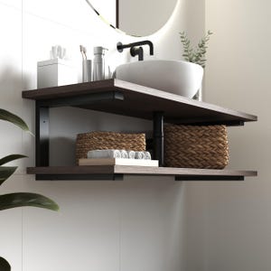 Puede incluir: Un mueble de baño montado en la pared con dos estantes de madera oscura sostenidos por soportes de metal negro. El estante superior tiene un lavabo blanco con un grifo negro. El estante inferior contiene dos cestas tejidas y toallas blancas.