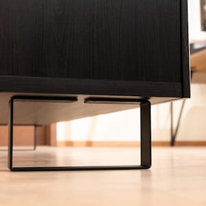 Puede incluir: Un mueble de madera negro con un acabado de grano oscuro y liso, apoyado por dos patas rectangulares de metal negro mate fijadas en la parte inferior. La pieza tiene un diseño lineal y limpio con un marco rectangular de tonos oscuros.