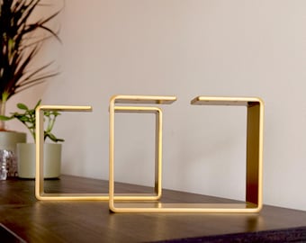 Könnte beinhalten: Drei goldene Tischbeine aus Metall mit quadratischer Form. Die Beine sind aus dickem Metall gefertigt und haben eine polierte Oberfläche.