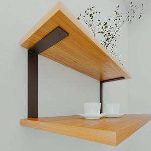 May include: Two-tiered wooden floating shelves with dark metal brackets. The shelves are made of light-colored wood and are mounted on a white wall. Two white teacups and saucers are on the lower shelf. Decorative sprigs are in the corner.