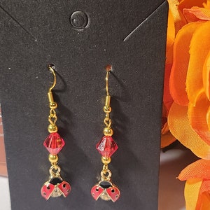 May include: A pair of gold dangle earrings with red and black ladybug charms. The earrings feature a red crystal bead and gold beads.