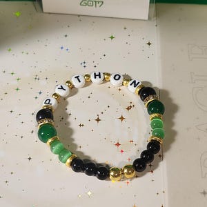 Può includere: Un braccialetto di perline con la parola "PYTHON" scritta con perline di lettere bianche. Il braccialetto presenta un mix di perline verdi, nere e dorate. Lo sfondo include una foto e una carta verde.