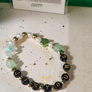 Puede incluir: Una pulsera de cuentas con el nombre "JINYOUNG" escrito en letras doradas sobre cuentas negras. La pulsera también presenta cuentas doradas, blancas y verdes, y una cuenta en forma de flor.