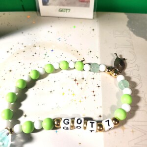 Puede incluir: Una pulsera de cuentas verdes, blancas y doradas. La pulsera incluye cuentas de letras blancas que deletrean "GOT7". Se adjunta un pequeño dije. Una tarjeta fotográfica con el texto "GOT7" está en el fondo.