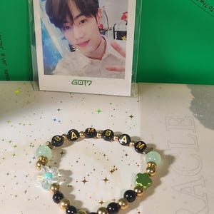 Może przedstawiać: Bransoletka z koralików z napisem "BAMBAM" zapisanym złotymi literami. Bransoletka ma czarne i złote koraliki, z jasnozielonymi i niebieskimi kwiatowymi akcentami. W tle zdjęcie osoby.