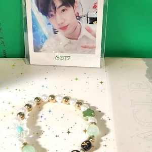 Może przedstawiać: Bransoletka z koralików z napisem "MAR" zapisanym czarnymi i złotymi koralikami. Bransoletka posiada również białe perły, złote koraliki oraz zielone i przezroczyste koraliki dekoracyjne. Karta ze zdjęciem z napisem "GOT7" znajduje się w tle.