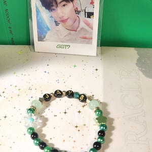 Op de afbeelding: Een kralenarmband met groene, zwarte en gouden kralen. De armband heeft een bloem- en klaverbedel, en de letters "JAYB" zijn gespeld in gouden en zwarte kralen. Een fotokaart met de tekst "GOT7" is op de achtergrond te zien.
