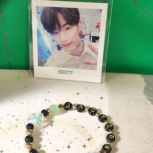Puede incluir: Una pulsera de cuentas con las letras "JACKSON" y cuentas en forma de estrella. La pulsera presenta cuentas negras, doradas y verdes, junto con una tarjeta fotográfica de una persona con el texto "GOT7". El fondo es una superficie verde.