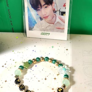 Peut inclure: Un bracelet de perles avec des perles de lettres noires épelant un mot, entrecoupées de perles dorées et vertes, et de breloques décoratives. Une photo d'une personne est derrière le bracelet, avec le texte "GOT7" en bas.