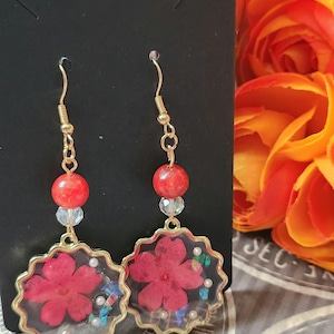 Può includere: Un paio di orecchini dorati con accenti di fiori rossi racchiusi in resina trasparente. Gli orecchini presentano una piccola perla rossa e un ciondolo di cristallo.