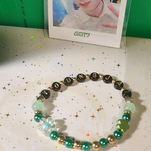 May include: A bracelet with green, gold, and black beads. The bracelet has the letters "JUNE" and a clear flower charm. A photo of a person is in the background, with the text "GOT7".