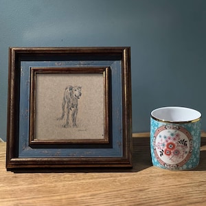 May include: A framed black and white pencil drawing of a dog standing in a field. The frame is blue with gold trim. The drawing is on a beige background.