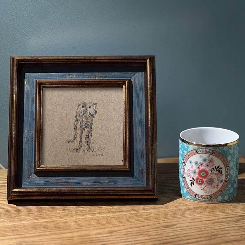 May include: A framed black and white pencil drawing of a dog standing in a field. The frame is blue with gold trim. The drawing is on a beige background.