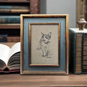 May include: A framed pencil sketch of a cat with a collar, set against a tan background. The frame has a distressed blue finish with gold trim. The artwork is displayed on a wooden surface.