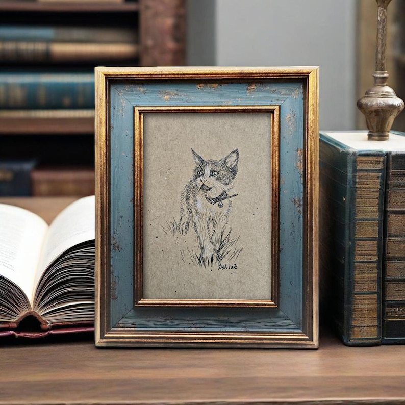 May include: A framed pencil sketch of a cat with a collar, set against a tan background. The frame has a distressed blue finish with gold trim. The artwork is displayed on a wooden surface.
