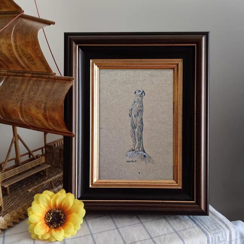 May include: A framed black and white drawing of a meerkat standing on a rock, with the artist's signature in the bottom right corner.