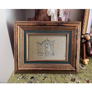 May include: A framed drawing of a cat with white fur and black markings. The cat is looking directly at the viewer. The frame is made of wood with a gold finish and a green inner border.