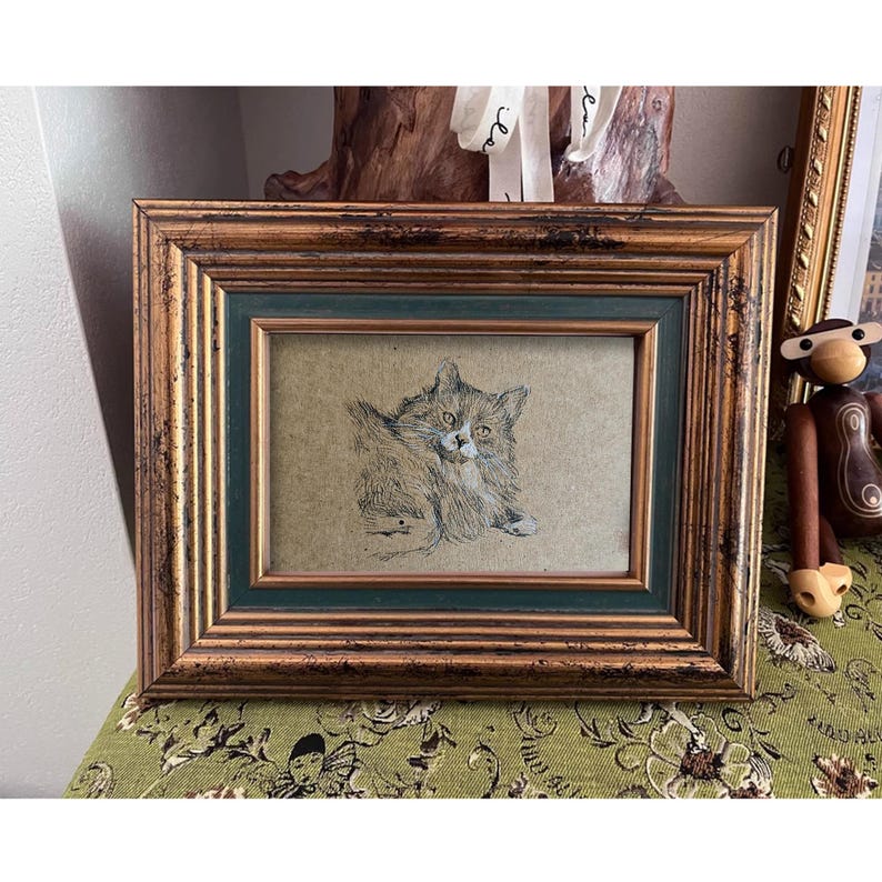 May include: A framed drawing of a cat with white fur and black markings. The cat is looking directly at the viewer. The frame is made of wood with a gold finish and a green inner border.
