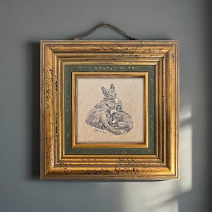 May include: A framed pen and ink drawing of two dogs, one lying on top of the other. The artwork is in a gold and green ornate frame, with a brown hanging cord. The drawing is signed "Tiger & Tom".