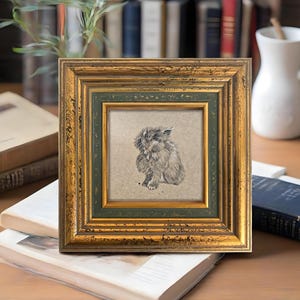 May include: Square framed artwork featuring a detailed pencil sketch of a fluffy rabbit. The frame has a gold finish with a green inner border. The artwork is displayed on a stack of books, suggesting a decorative home accent.