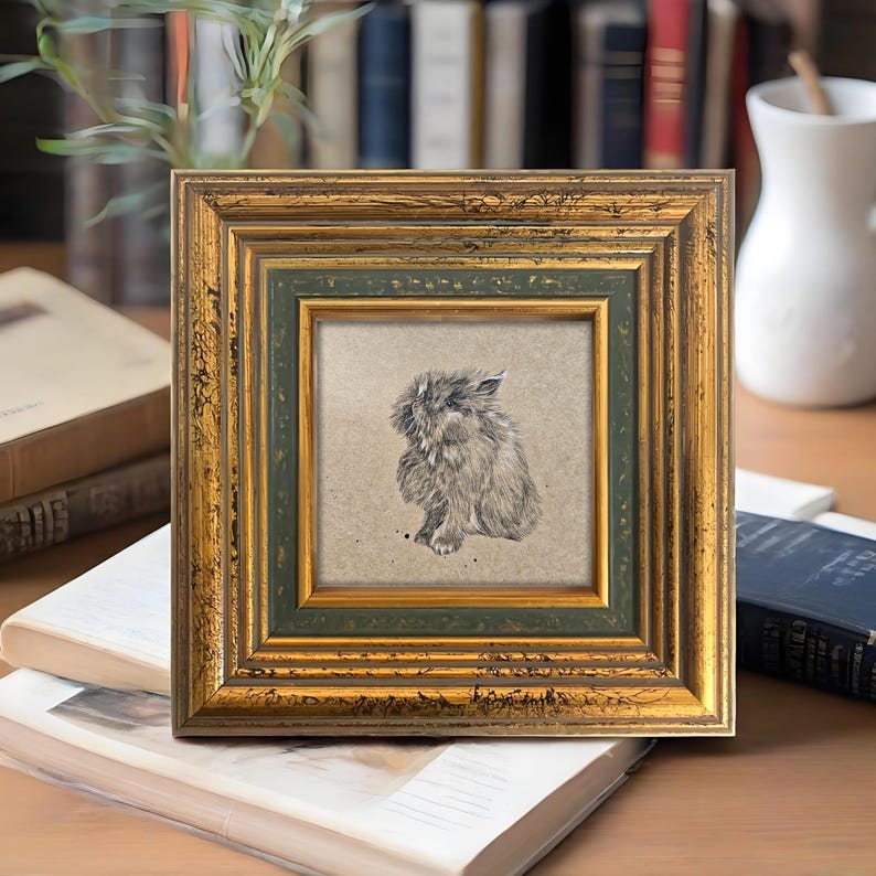 May include: Square framed artwork featuring a detailed pencil sketch of a fluffy rabbit. The frame has a gold finish with a green inner border. The artwork is displayed on a stack of books, suggesting a decorative home accent.
