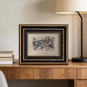 May include: A framed black and gold picture of a house, resting on a wooden surface. The picture is a black and white sketch of a house with the text "1969 John & Susan, Oregon" at the bottom. A lamp is to the right of the picture.