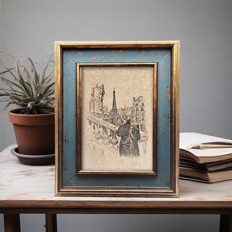 May include: A framed sketch of the Eiffel Tower and a person on a balcony. The frame has a distressed blue finish with gold trim. The artwork is displayed on a wooden table with a potted plant and a stack of books.