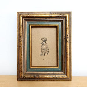 May include: A framed dog portrait. The frame has a gold and teal finish with a distressed look. The dog illustration is in black and white, set against a tan background. The frame is rectangular and has a double border.
