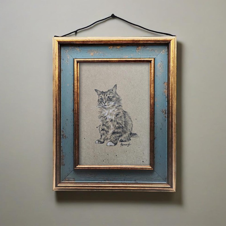 May include: A framed pencil drawing of a cat. The artwork is in a distressed blue and gold frame, with a black hanging cord. The cat is depicted in a seated position, with detailed fur and a focused gaze. The artwork is signed "Reany".
