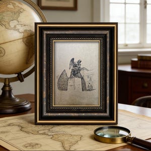 May include: A framed sketch of a statue with an angel figure, set in a black and gold ornate frame. The frame is displayed on a table with a vintage globe, a map, and a magnifying glass.