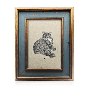 Op de afbeelding: Een ingelijste kattenkunstprint. De lijst heeft een distressed blauwe buitenkant met een gouden binnenrand. Het kunstwerk is een gedetailleerde zwart-wit illustratie van een tabby kat die zichzelf verzorgt.